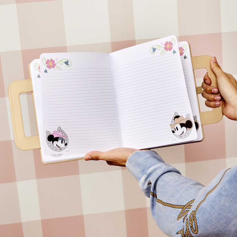 Someone holding open the Loungefly Western Mickey and Minnie Lunchbox Stationery Journal to show off the lined pages with western Mickey and Minnie in the bottom corners of the pages and floral designs in the upper corners against a pink plaid background 